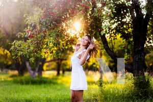 A person in a white dress stands among blooming trees at sunset in a lush garden, with golden sunlight filtering through leaves.