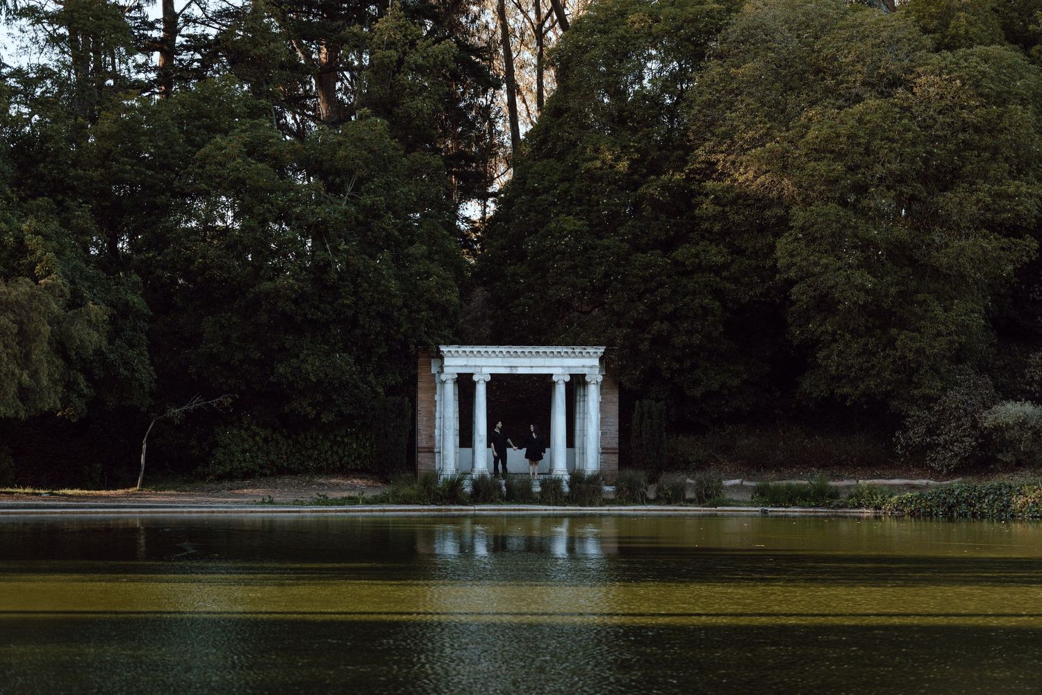 A classical white gazebo reflects in still water surrounded by trees at dusk.