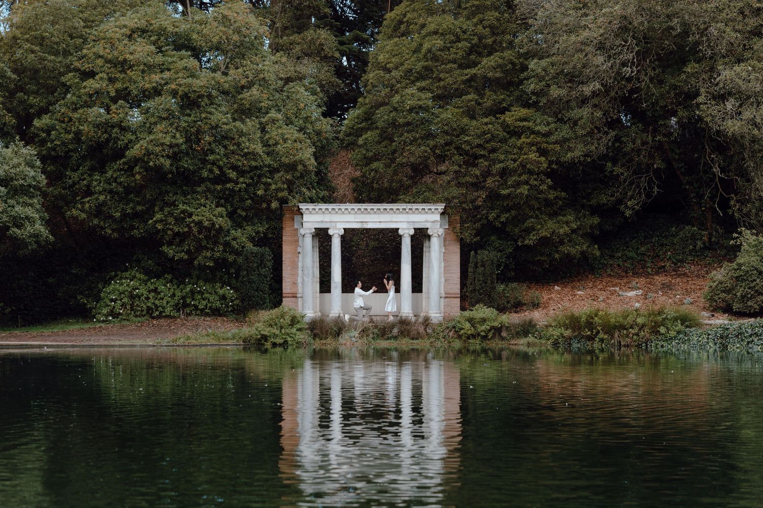 A classical white gazebo reflects in still water surrounded by trees at dusk.