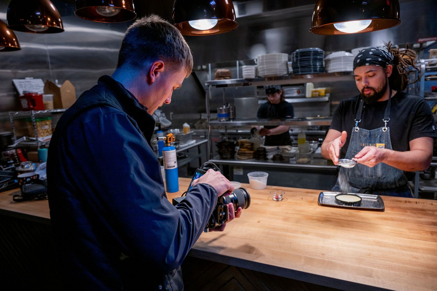 Two chefs preparing dishes in a modern restaurant kitchen under warm copper pendant lights.