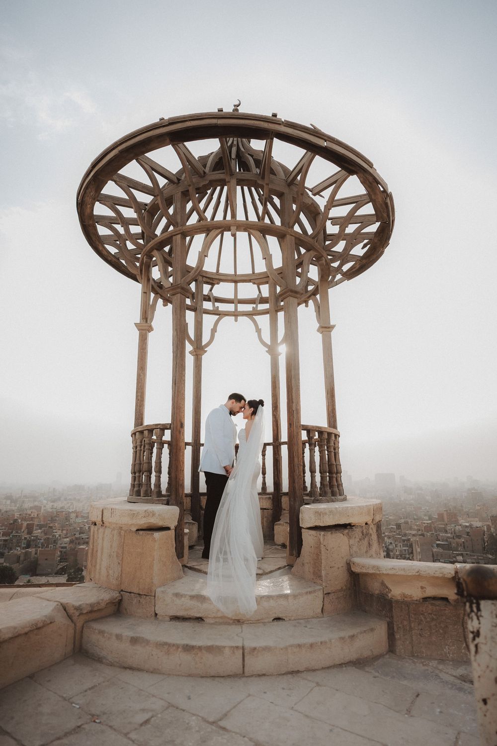 A couple embraces under an ornate vintage gazebo with decorative metalwork dome against a misty cityscape backdrop.