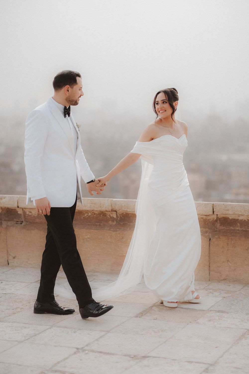 A bride in a flowing white gown and groom in white jacket share a romantic moment on a misty balcony.