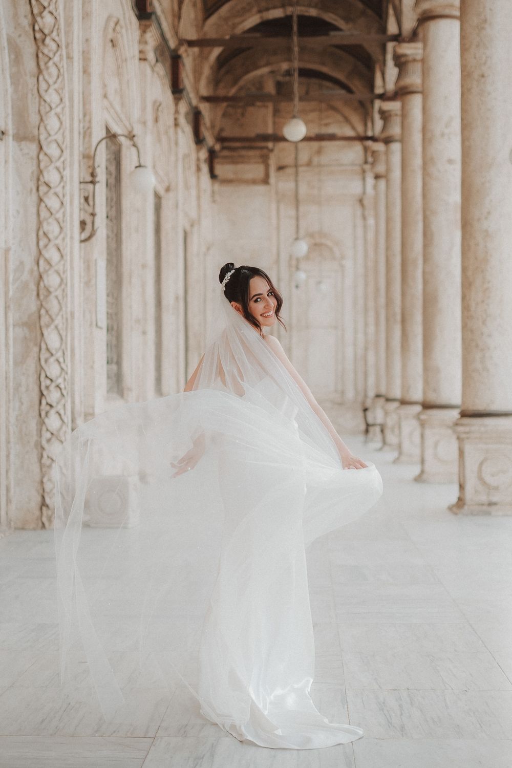 A bride in a flowing white gown and veil twirls elegantly beneath an ornate arched colonnade.