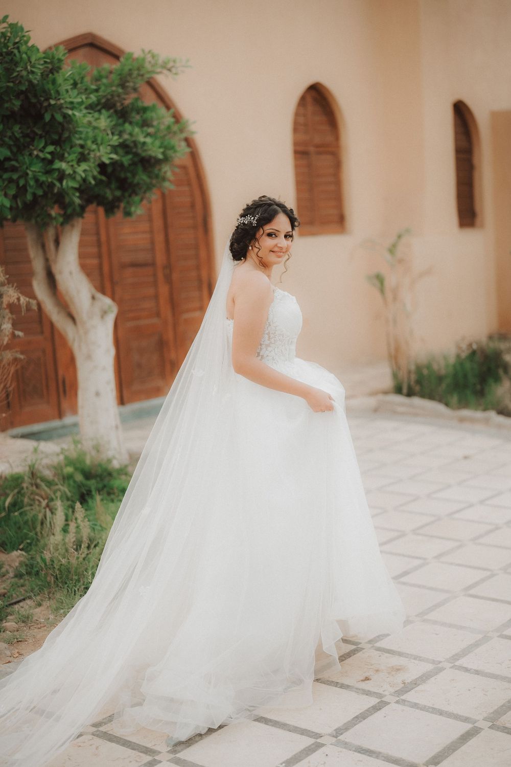 A bride in a flowing white ball gown and veil poses gracefully against a Mediterranean-style building with arched doorways.