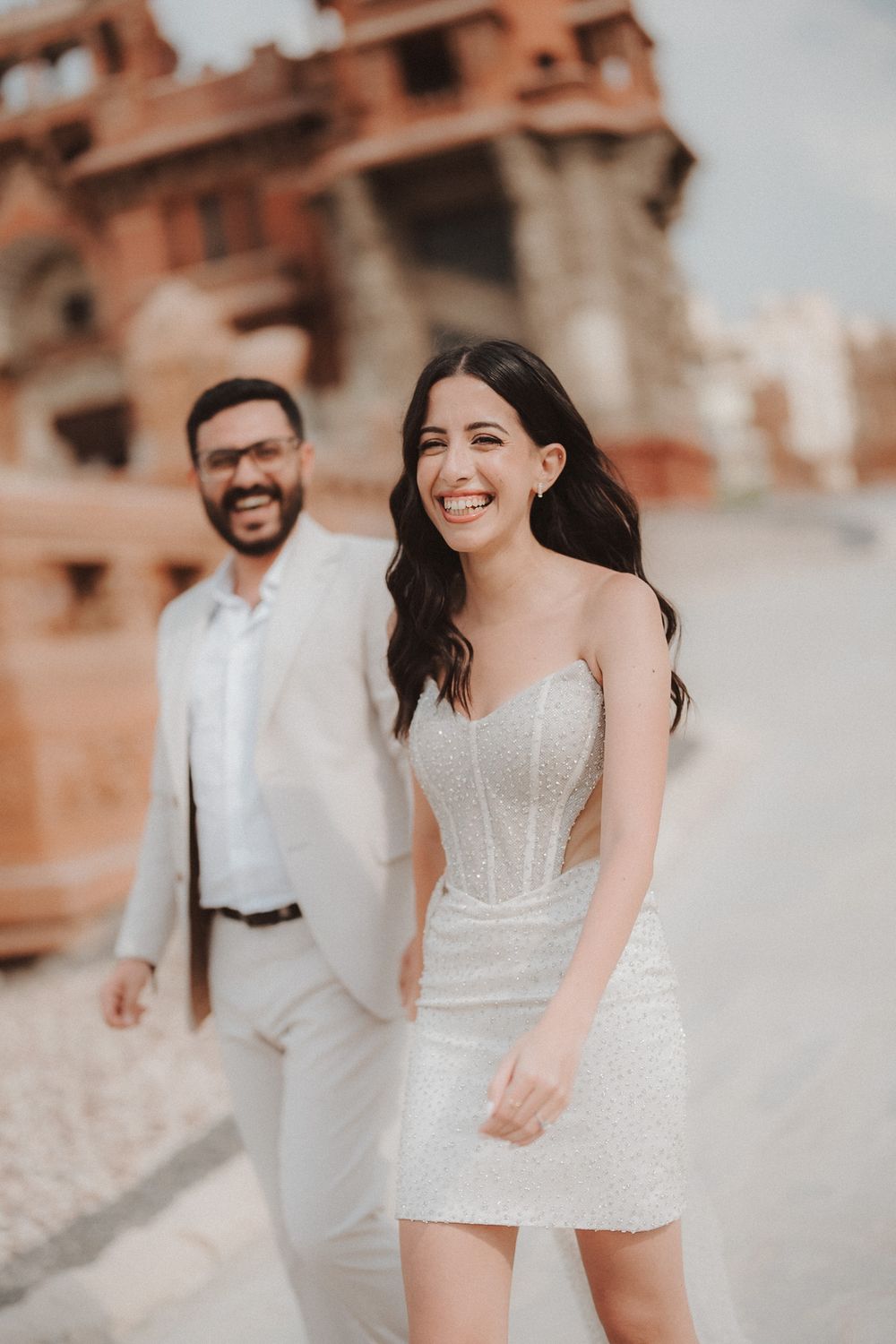 A couple laughs together during a casual photoshoot against a rustic terracotta building backdrop.