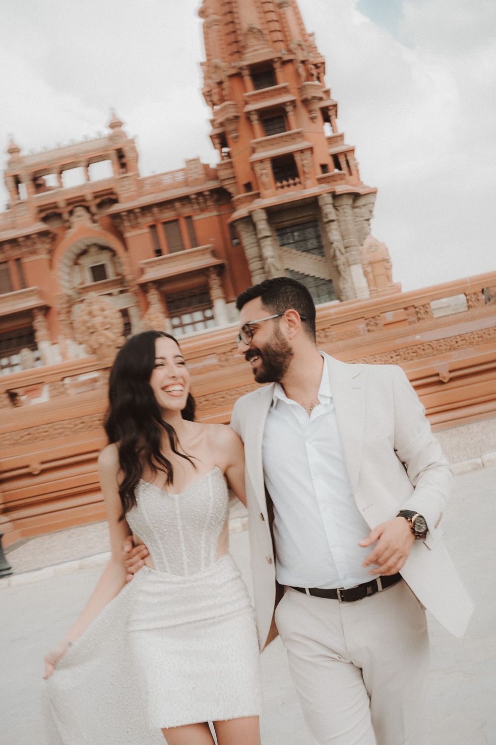 A couple in matching white outfits share a romantic moment in front of an ornate terracotta-colored palace building.