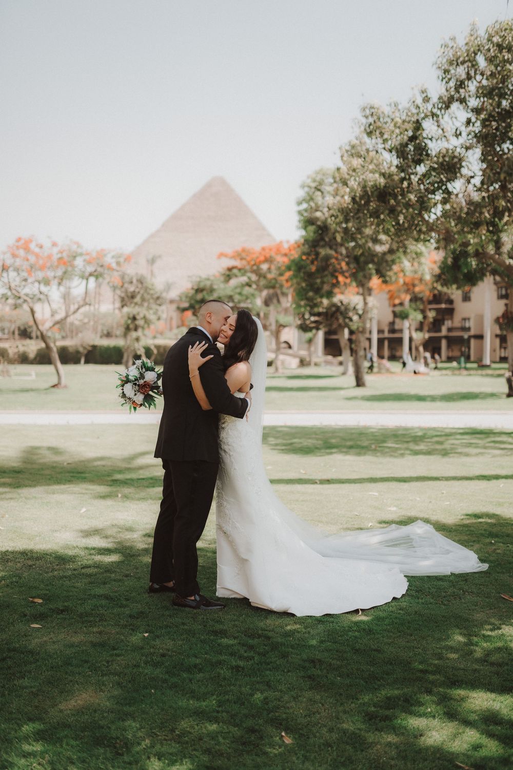A couple in formal wedding attire dips and embraces on a grassy lawn with buildings in the background.