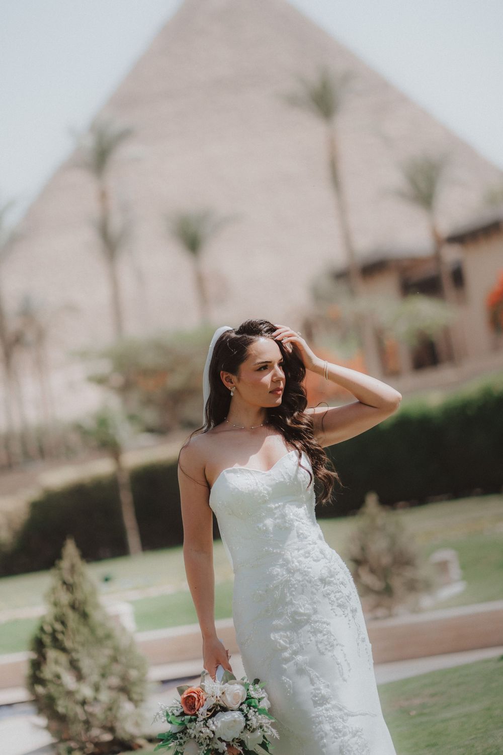 A bride in a white lace dress poses elegantly outdoors with palm trees in the background.