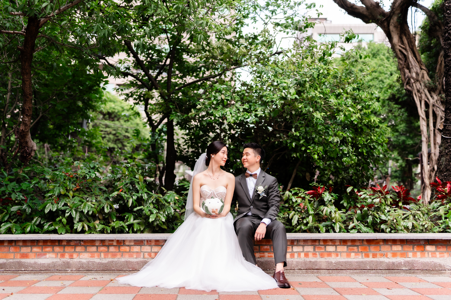Couple posing on garden bench surrounded by lush greenery and brick wall.
