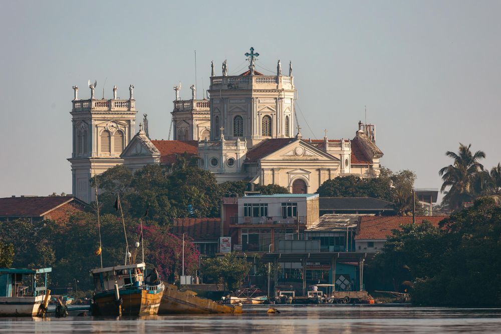 Historic white colonial church with twin towers overlooks waterfront buildings in Sri Lanka during sunrise.