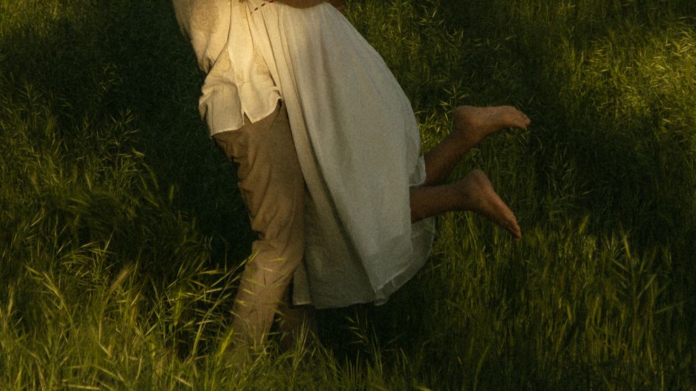 Two people in casual attire embrace in tall grass during golden hour. Documentary photographer
