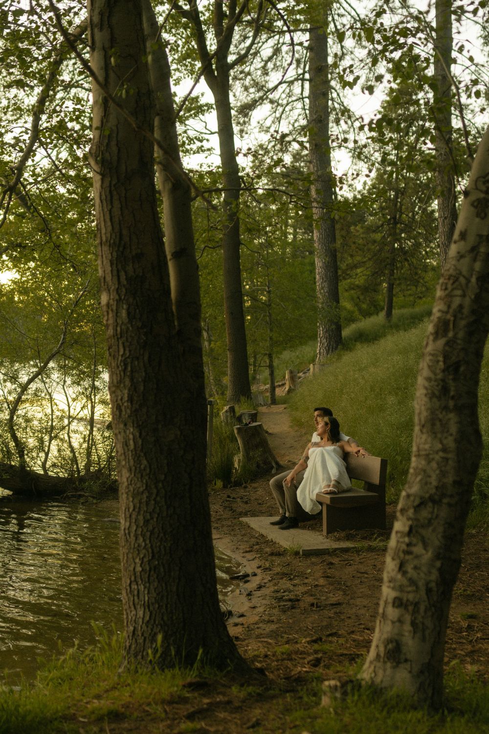 A couple in white sits on a bench along a tree-lined waterfront path at sunset. Documentary photographer
