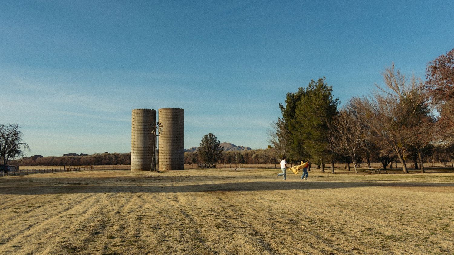 Two tall cylindrical concrete silos stand in a dry winter field under blue sky with bare trees in the background.