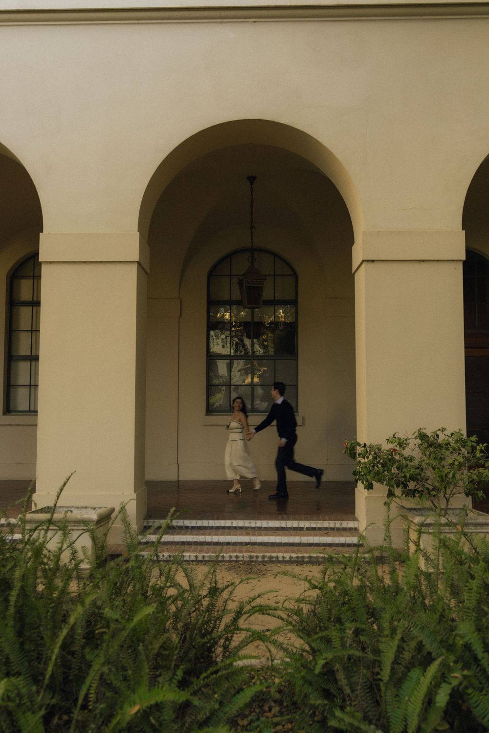 Couple running through arched colonnade with garden landscaping in foreground. Documentary photographer