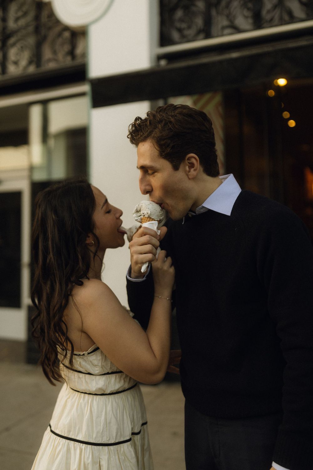 Couple sharing ice cream cone outside storefront in evening light. Documentary photographer