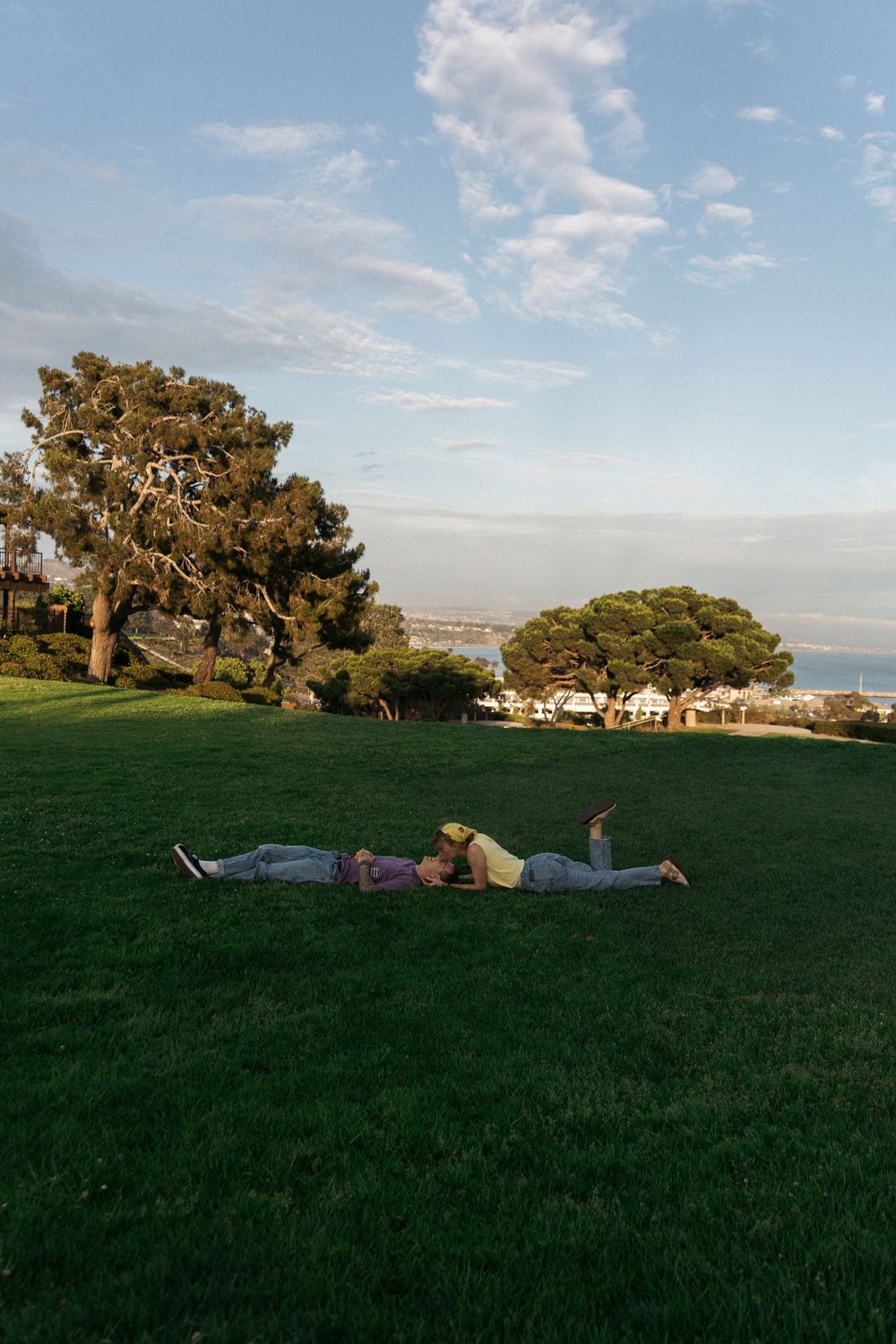 People relaxing on a grassy hill overlooking a coastal town with trees and blue sky on a sunny afternoon.