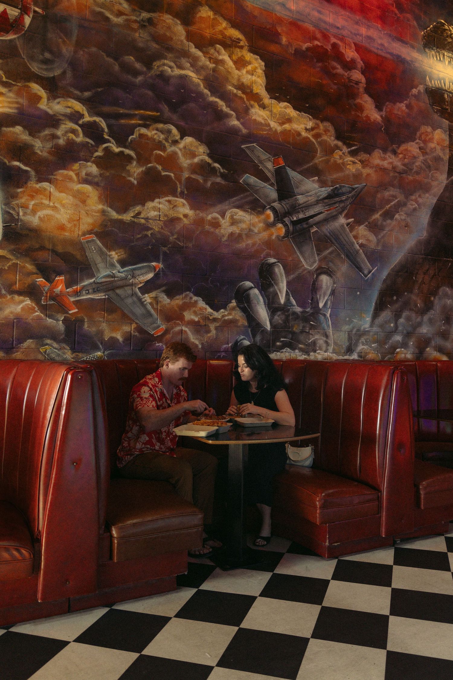 Couple having lunch at Retro diner booth with red leather seats beneath dramatic mural of fighter jets in stormy clouds. Documentary photos