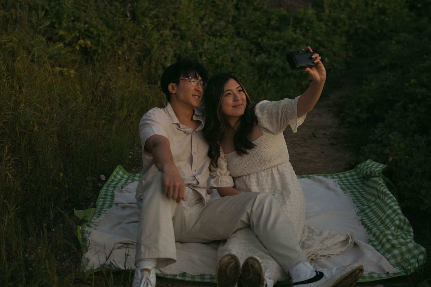A couple in white outfits takes a selfie while sitting on a blanket outdoors. Film camera Documentary photographer
