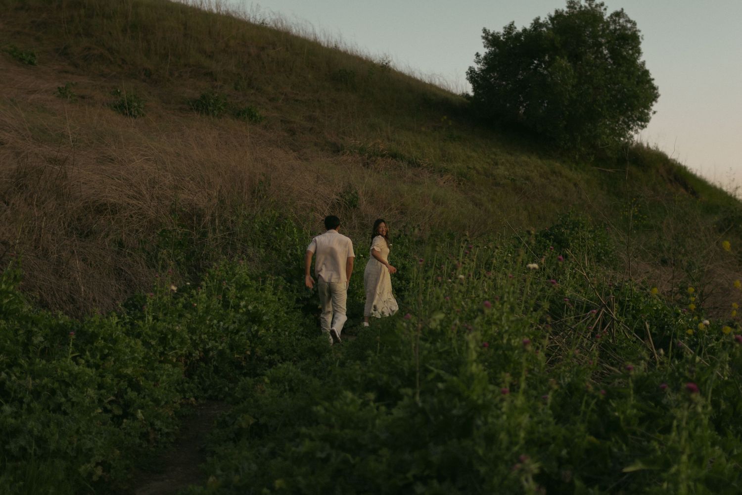 Two people in light-colored clothing walk through tall grass on a hillside at dusk. Documentary photographer