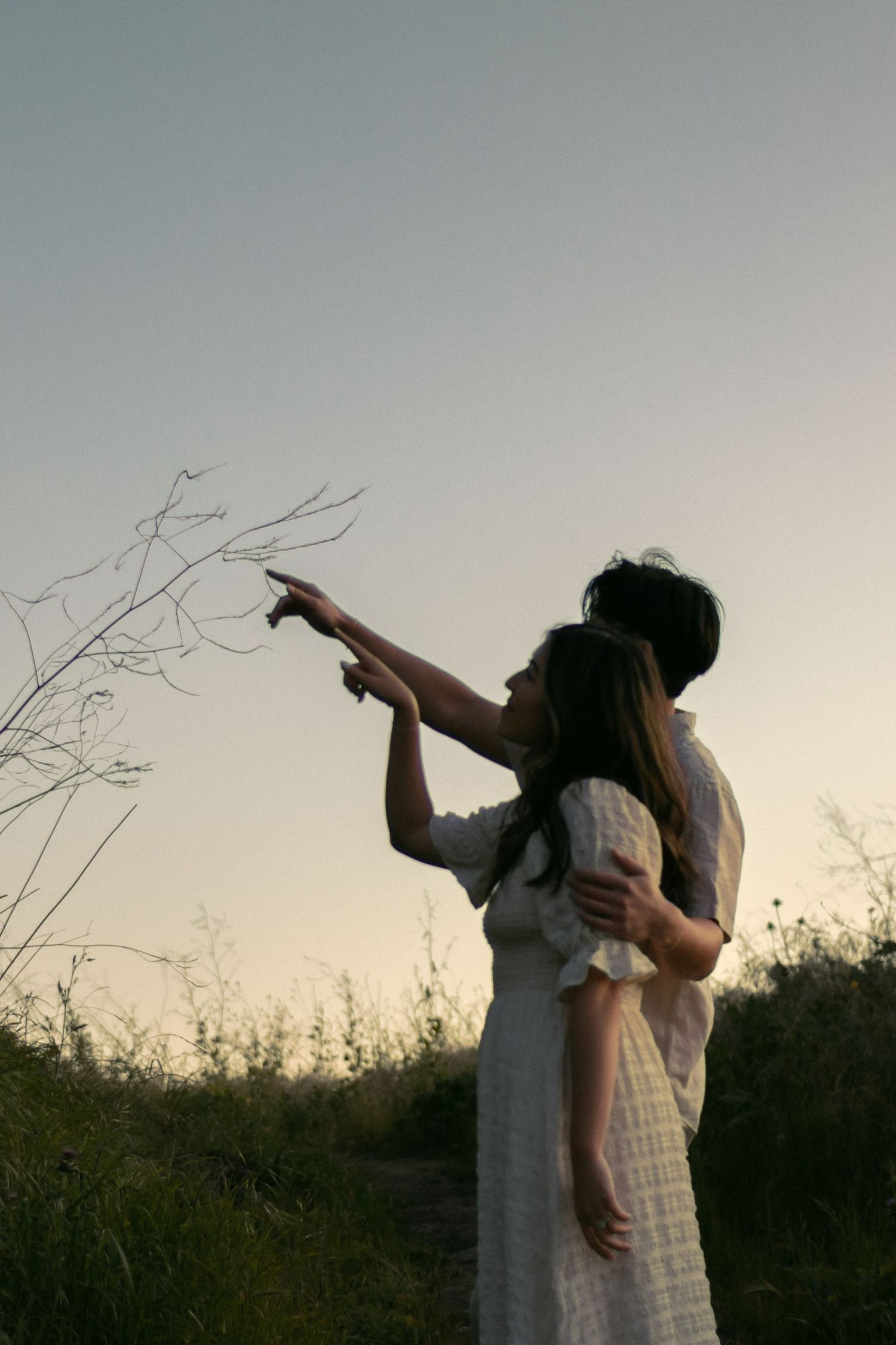 A couple in white dresses points at the sky during sunset in a grassy field. Documentary photographer