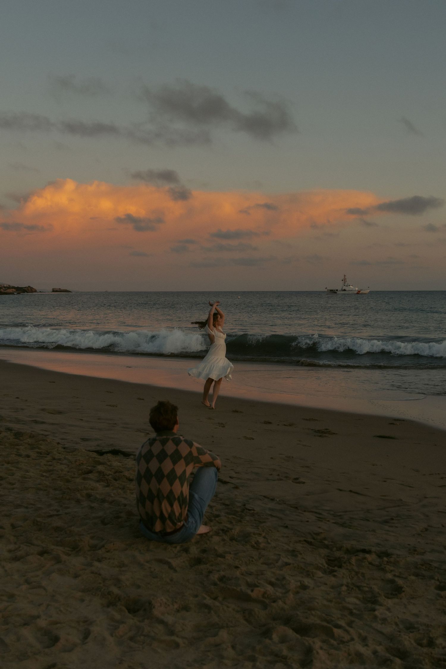 couple share a romantic moment on the beach at sunset with a ship visible on the horizon. with natural documentary style photography.