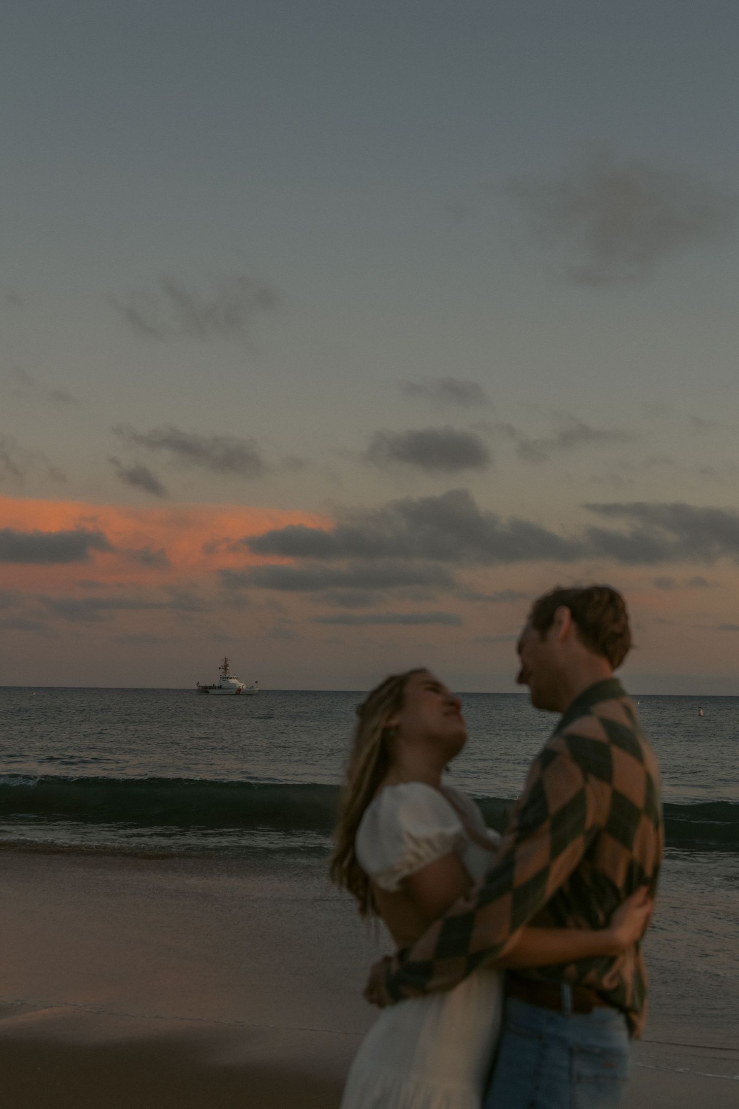 A couple embraces on a beach during golden hour with a boat in the distance. with natural documentary style photography. Beach session