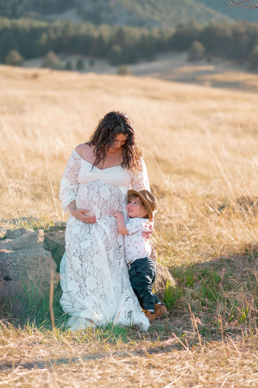 Pregnant woman in white lace gown with toddler in cowboy hat embracing her bump in golden field.