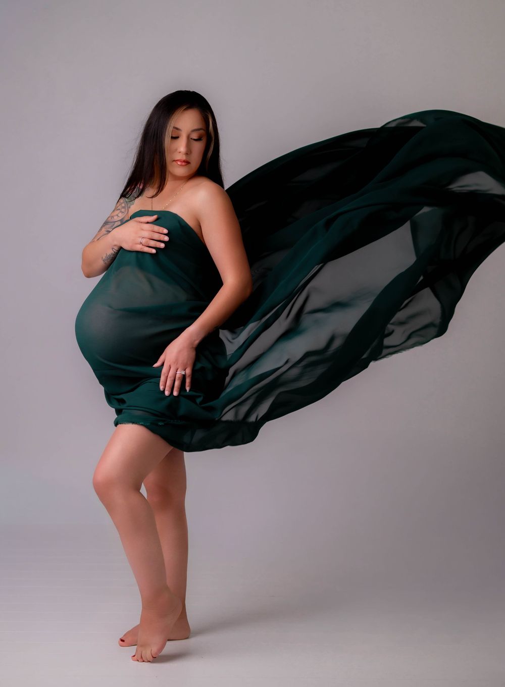 Pregnant woman draped in flowing emerald green fabric poses barefoot in a studio maternity photography session.