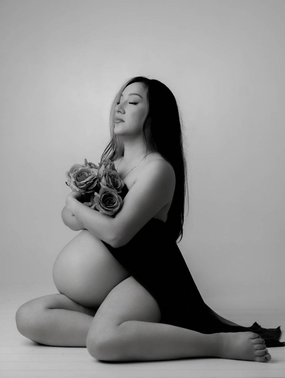 Black and white maternity photo of a pregnant woman sitting gracefully, holding roses, with long dark hair.
