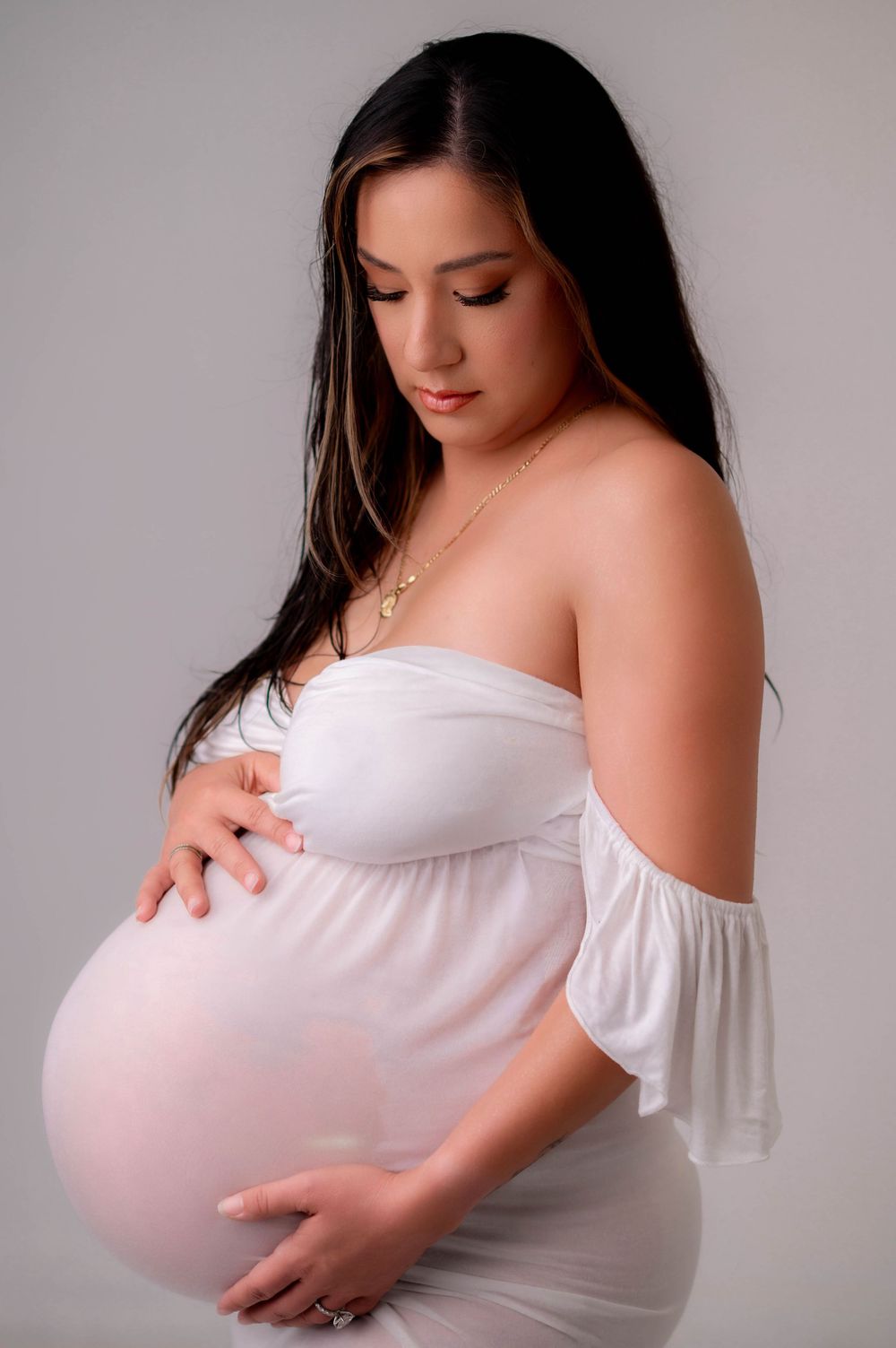 Pregnant woman in white off-shoulder dress cradling baby bump, wearing gold necklace, studio maternity portrait.