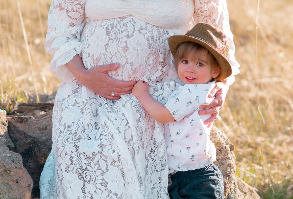 A toddler in a hat hugs a pregnant woman in a white lace dress outdoors in a golden field.