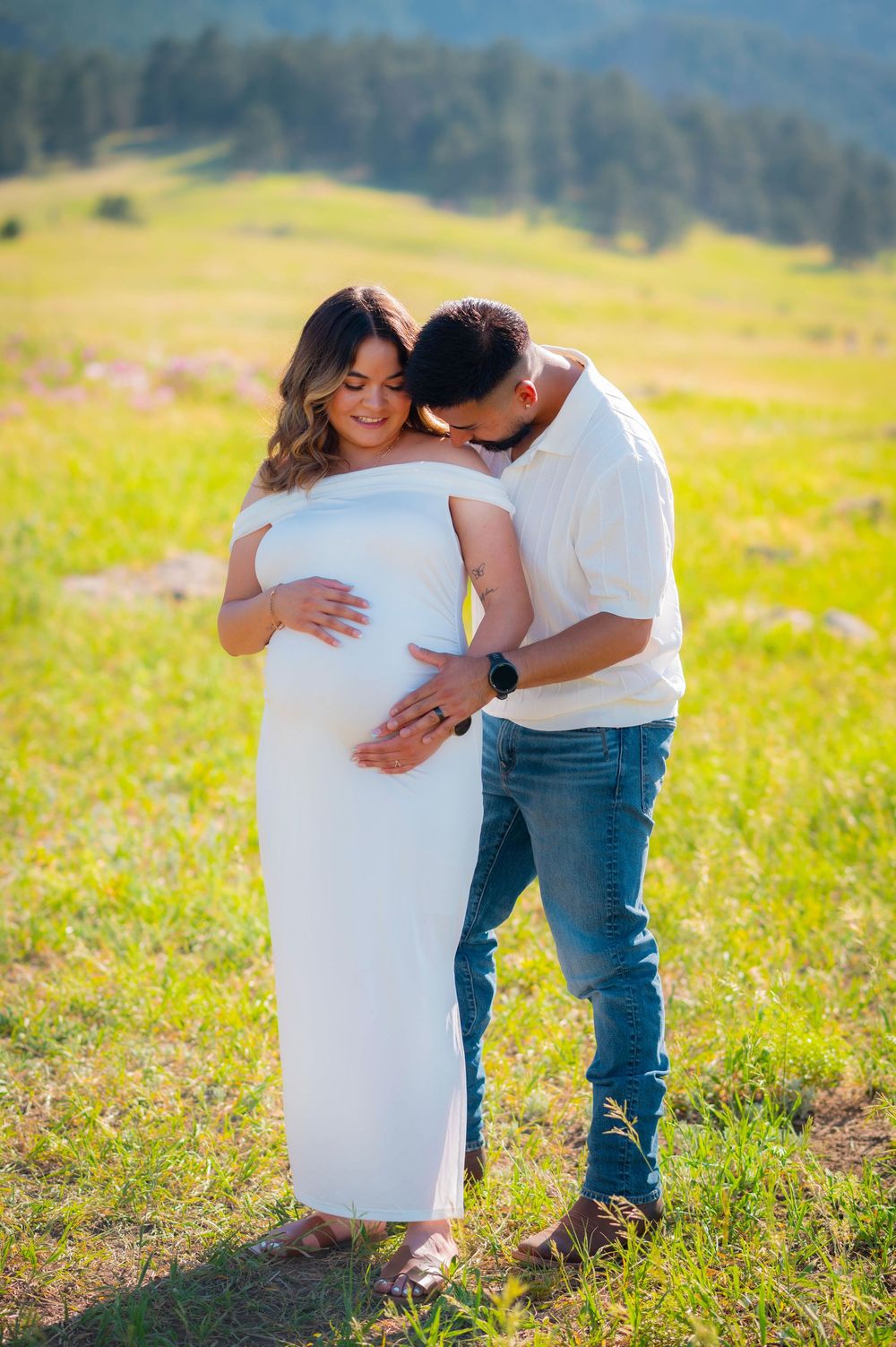 Couple shares tender maternity photo outdoors, woman in white dress cradling baby bump while partner kisses her head in green field.