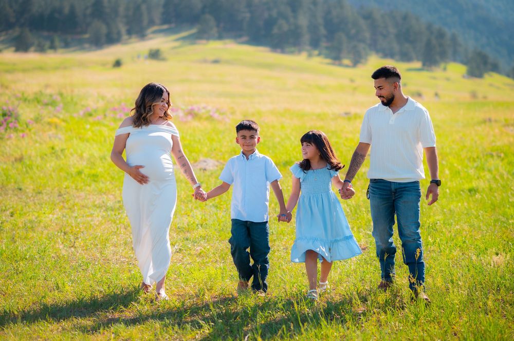 Pregnant woman and man walk with two children in a sunny green meadow, all wearing white and blue outfits.