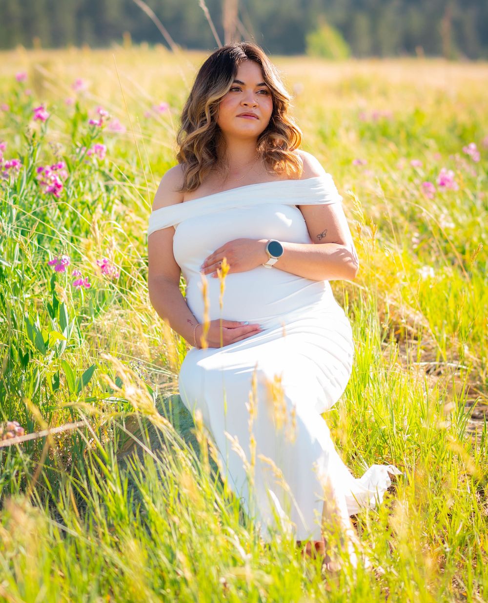 Pregnant woman in white off-shoulder gown posing in a sunlit wildflower meadow during a maternity photoshoot.