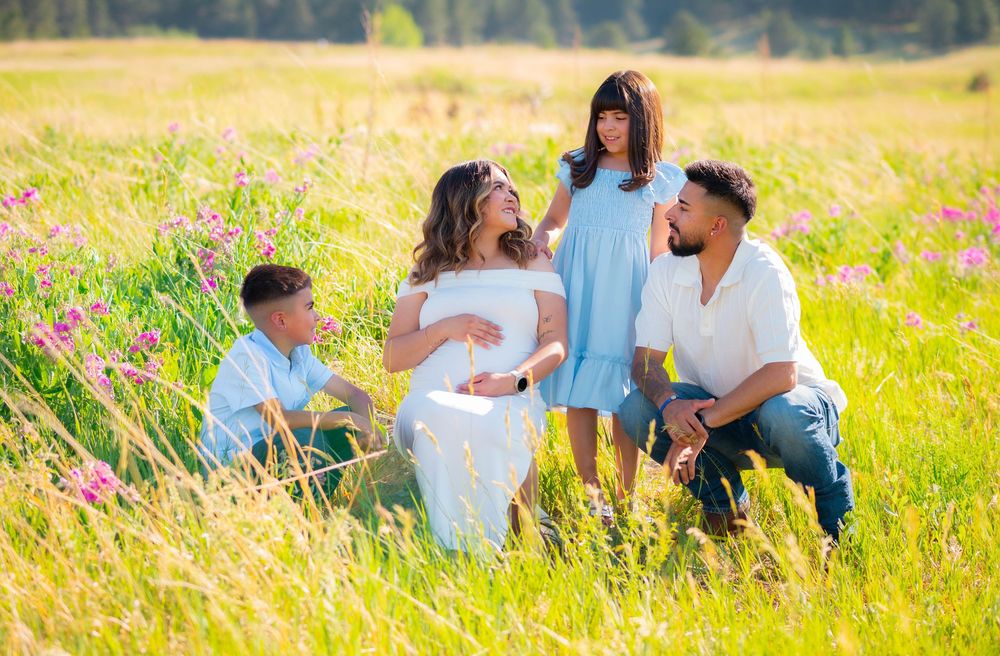 Pregnant woman in white dress with family in sunny wildflower meadow, maternity photo session outdoors.