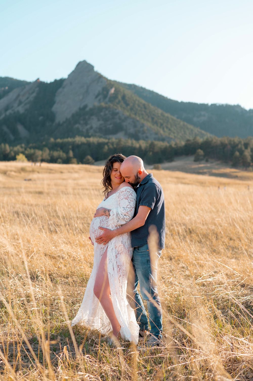 Pregnant couple embracing in golden field with Rocky Mountain Flatirons backdrop during maternity photo session.