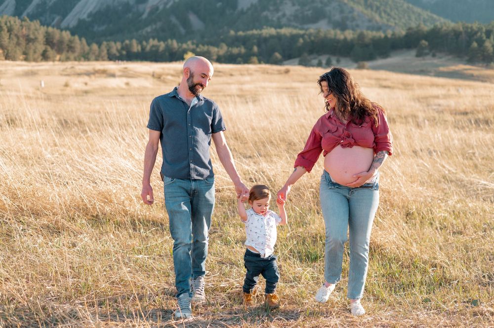 Pregnant couple holding toddler's hands while walking through golden meadow with mountain backdrop.