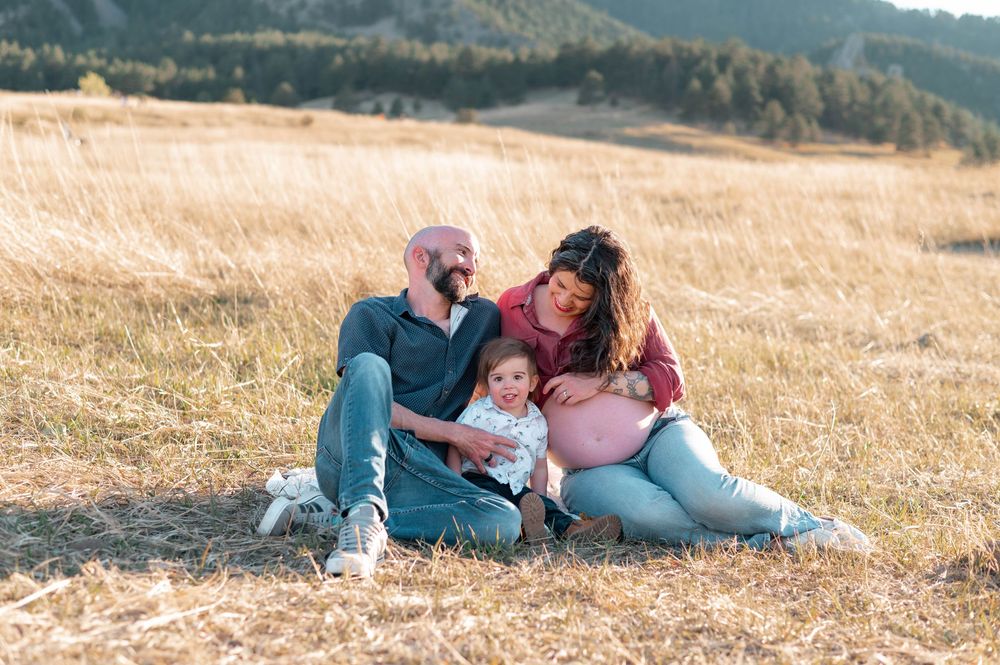 Pregnant family sitting in golden field with toddler, mountains in background, maternity photo session outdoors.