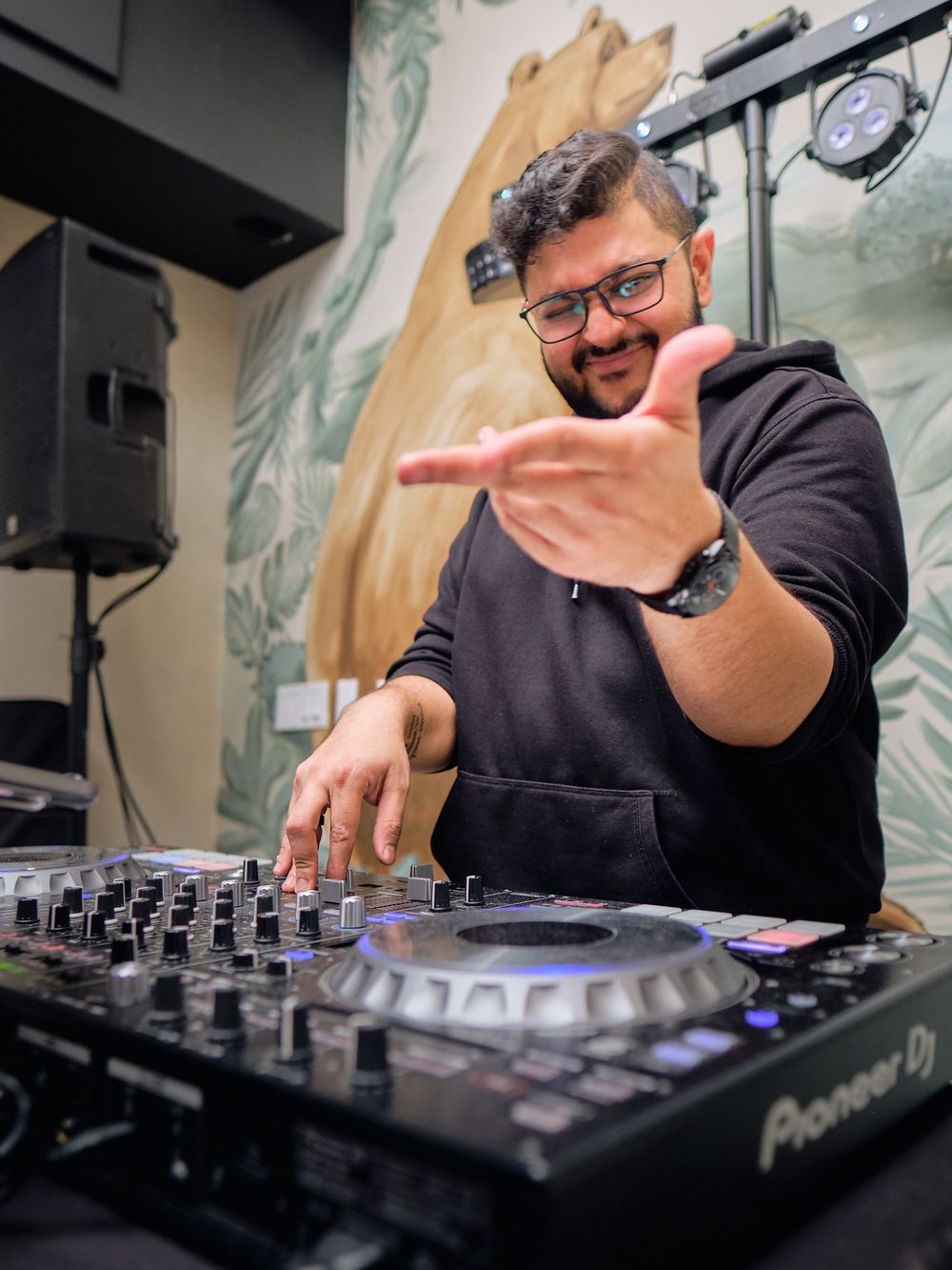 DJ performs behind Pioneer equipment while wearing a black hoodie and gesturing at a music venue with decorative wall art.