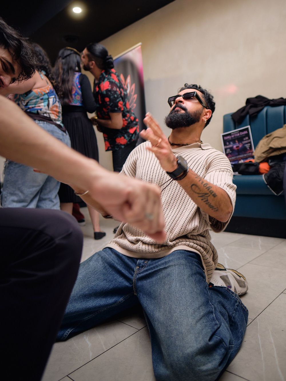 Someone in sunglasses and striped sweater sits on floor at party while reaching for something with dramatic expression.