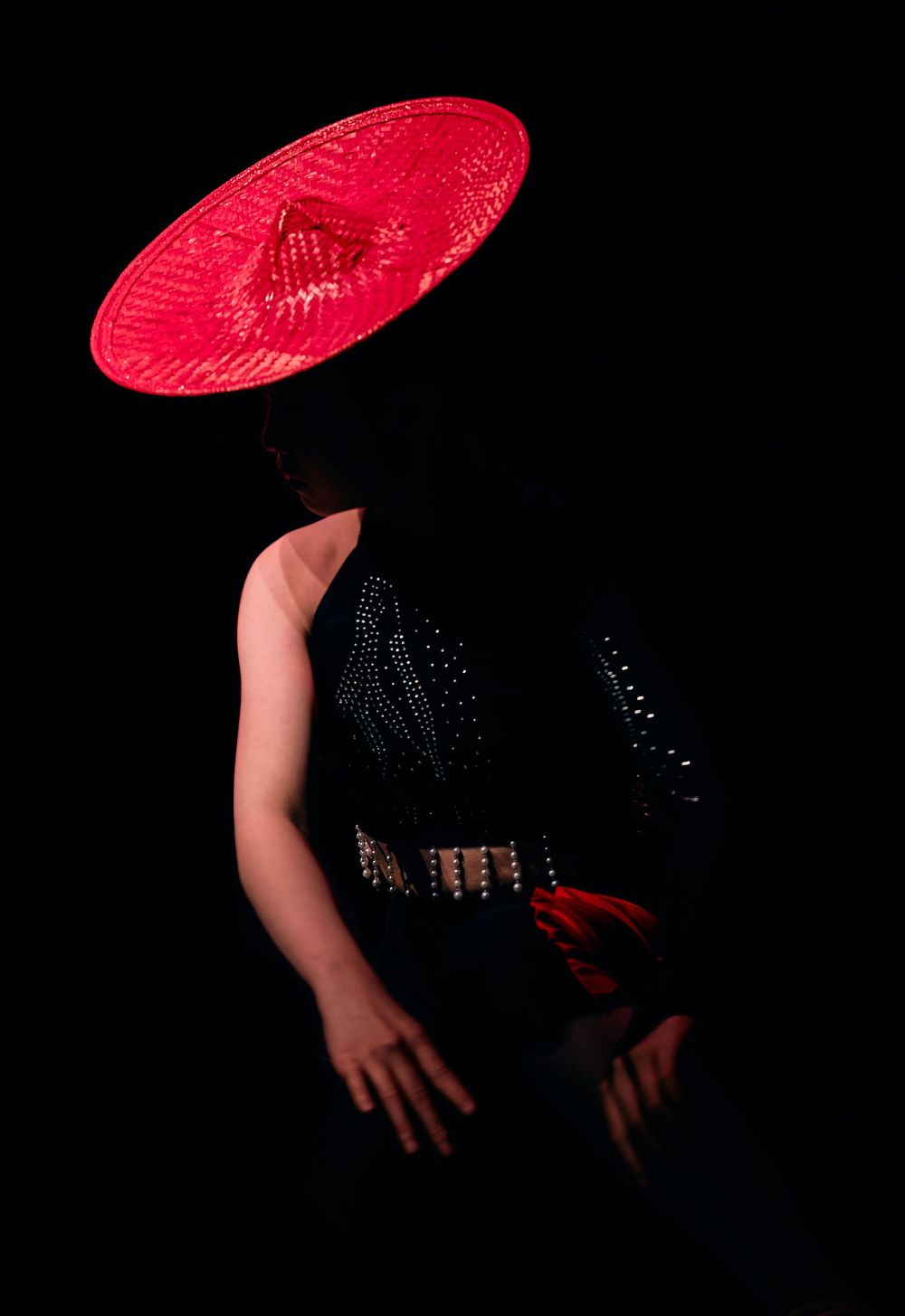 Dramatic silhouette wearing black dress and red circular hat against dark background creates striking contrast.