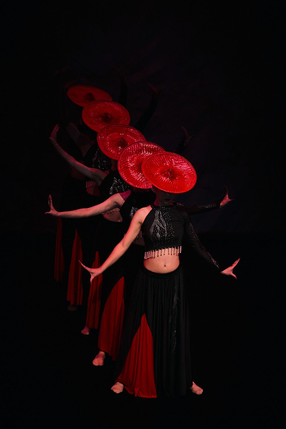 Multiple dancers in black outfits with red parasols performing against dark backdrop.