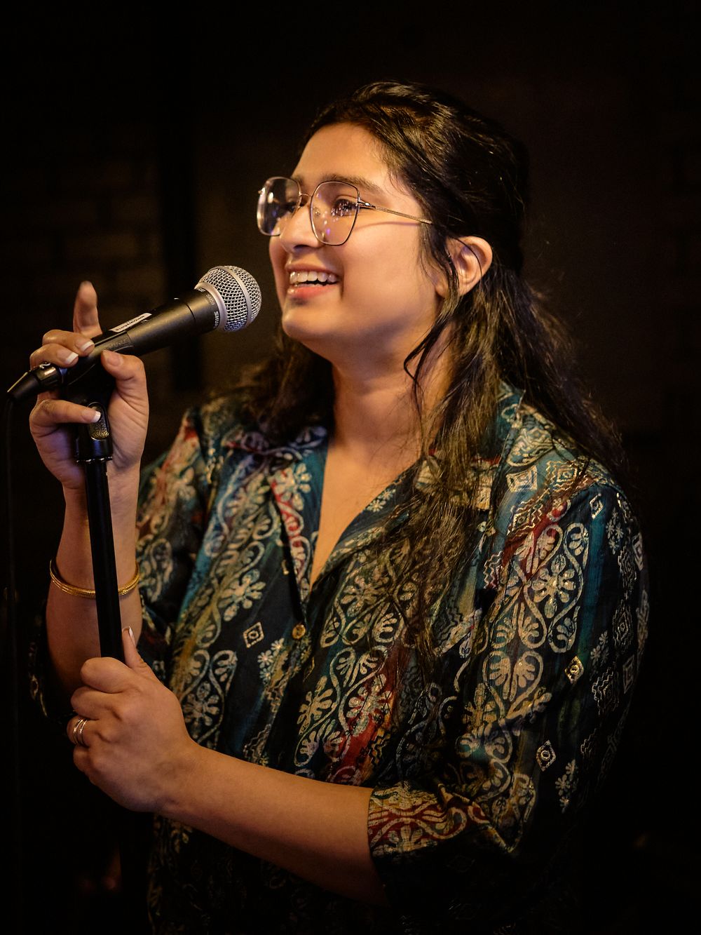 A performer in glasses and a patterned blue shirt smiles while speaking into a microphone on a darkened stage.