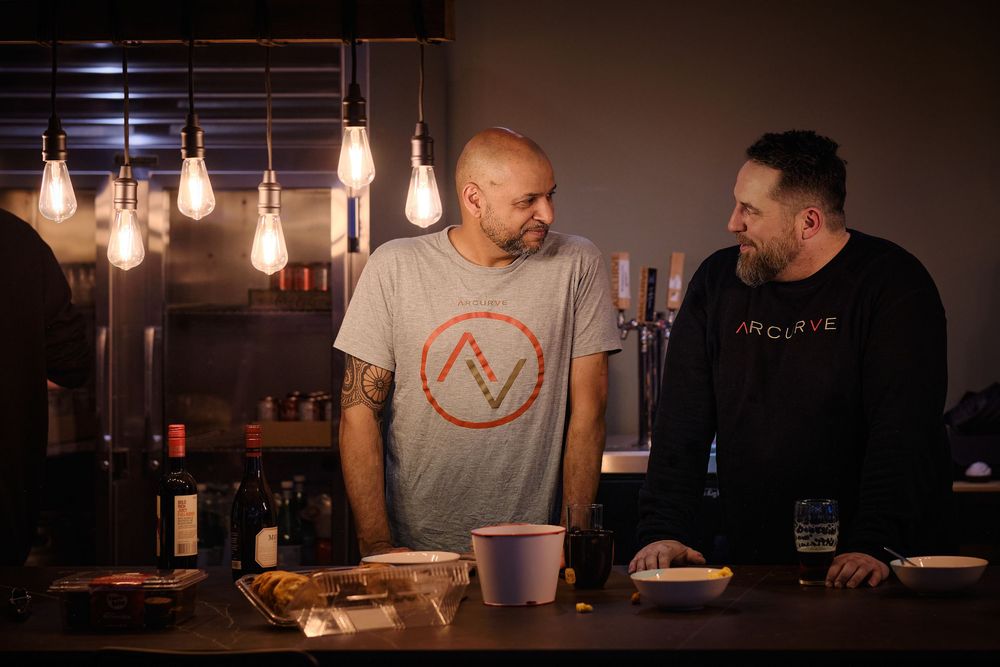 Edison bulbs illuminate a dark bar interior while two people converse at the counter.