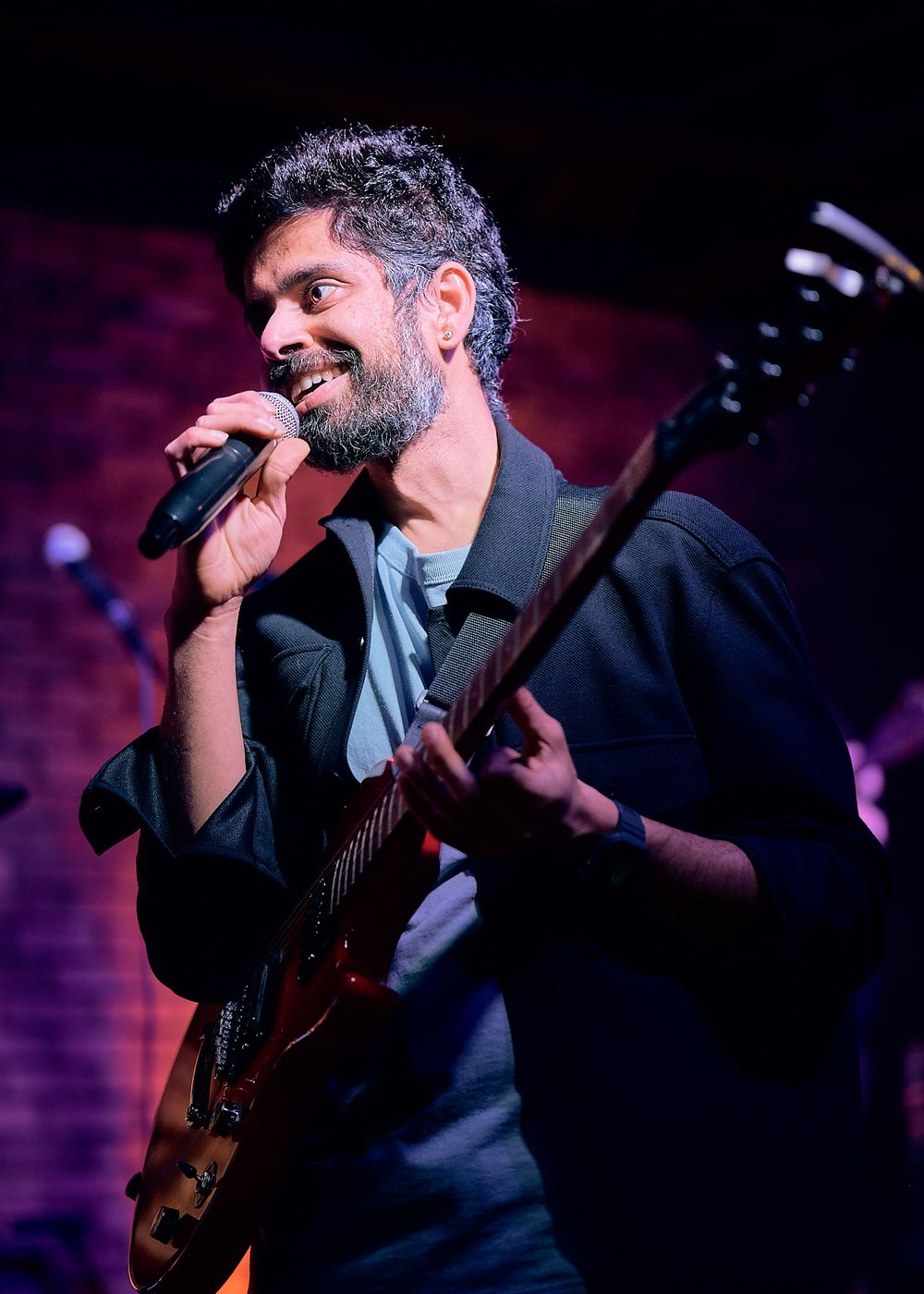 A performer singing with emotion while playing an electric guitar on stage with dramatic purple lighting in the background.