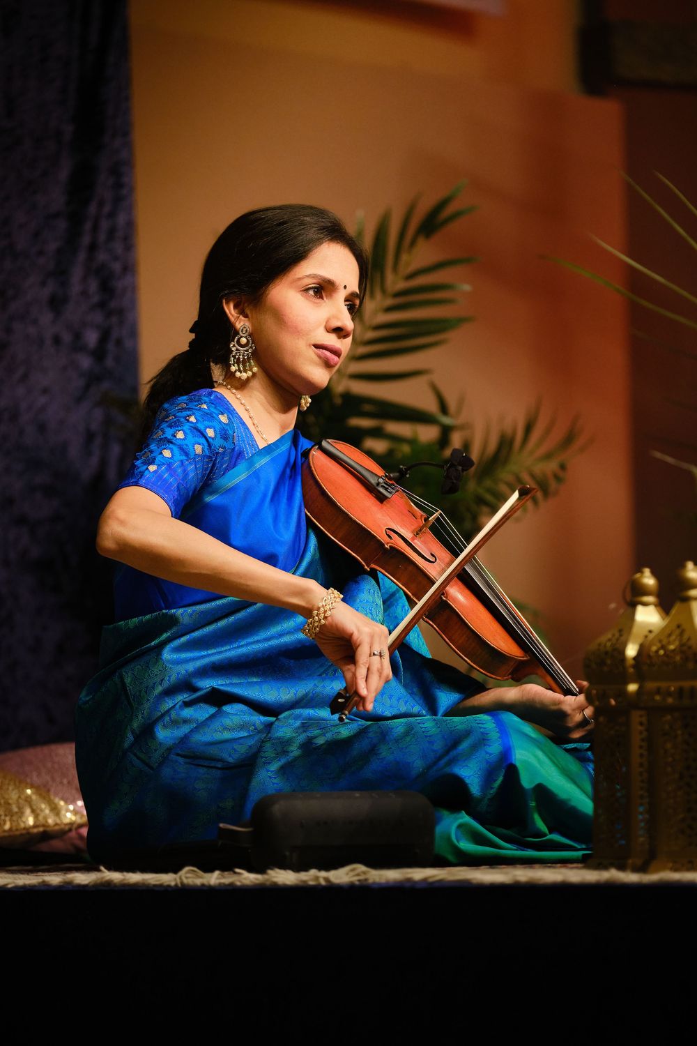 A musician in a blue and teal sari performs on violin while seated on stage with palm decor in the background.