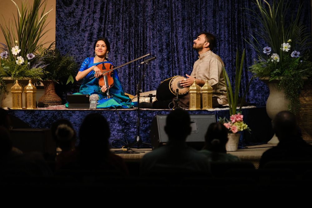 Musicians perform on stage with blue curtain backdrop and decorative plants during an evening concert.