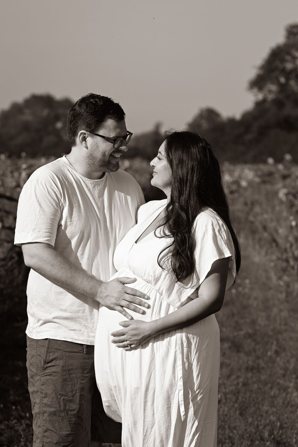 A couple in white clothing share an intimate moment outdoors while expecting a baby in this black and white portrait.