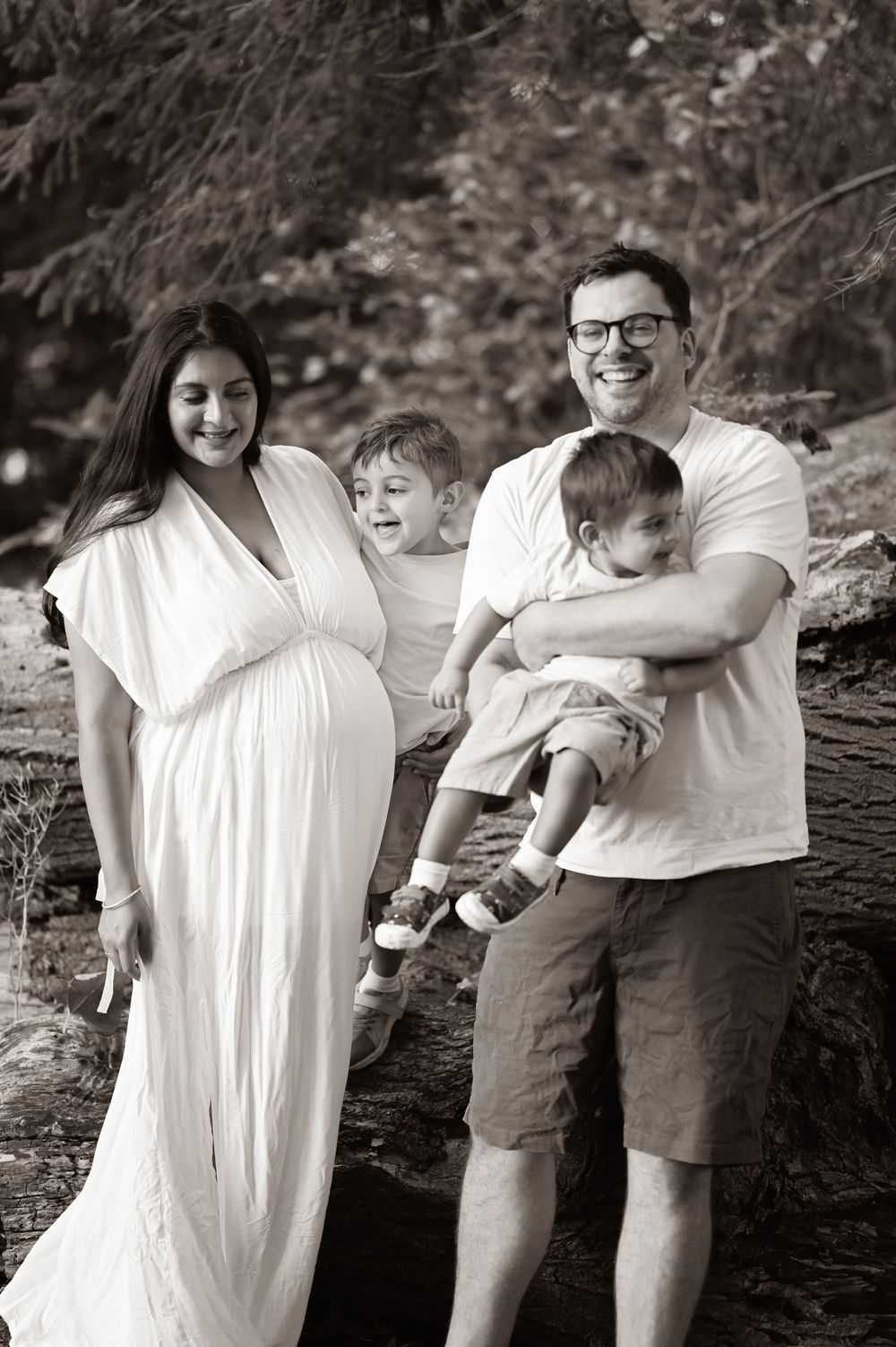 A black and white portrait of a family in a natural outdoor setting with a pregnant woman in a white dress.