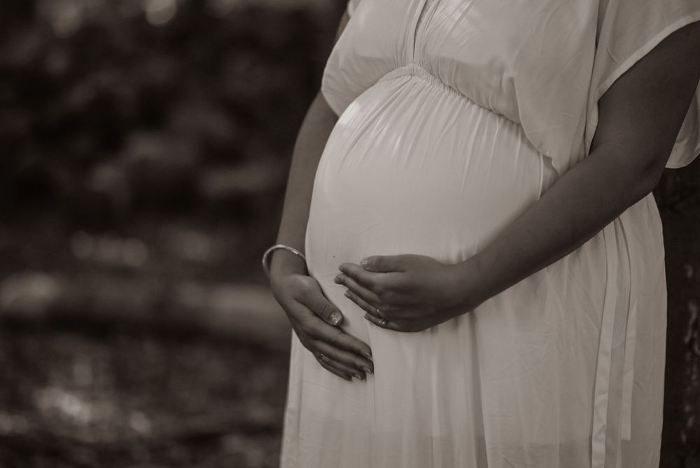 Close-up black and white image of a pregnant belly in a flowing white dress with hands gently cradling the bump.